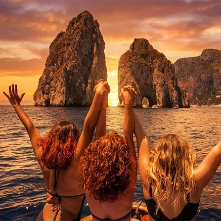 Three female friends celebrating on a private boat charter during a luxury Capri sunset cruise with the Faraglioni rocks in the background.